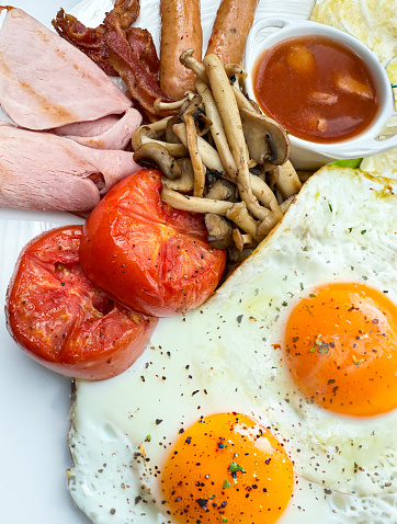 Stock photo showing close-up, elevated view of fried breakfast on white plate, slice of white toast, two sunny-side-up fried eggs, ramekin of baked beans, sausages, streaky bacon rashers, ham slices, grilled tomatoes and mushrooms.