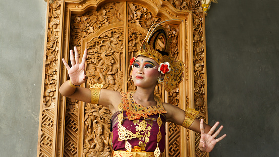 balinese girl dance gesture wearing Balinese traditional dress with a dancing gesture with crown, jewelry, and gold ornament accessories on doors carved with Balinese motifs background