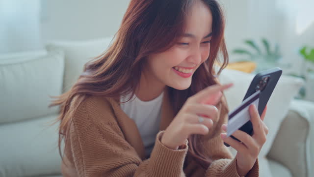 Happy young asian woman holding credit card using instant mobile payments at home. Smiling Female customer shopper making purchase on smartphone receiving cash back concept. E-banking app service.