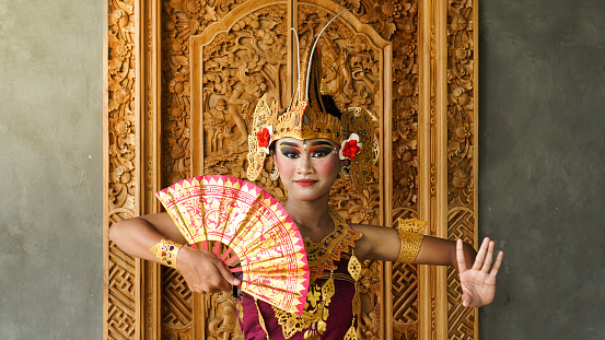 balinese girl dance gesture wearing Balinese traditional dress with a dancing gesture with crown, jewelry, and gold ornament accessories on doors carved with Balinese motifs background