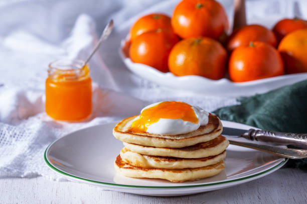 Pancakes served with tangerine jam and sour cream, and tangerines on a light background.