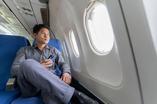 Young professional Asian man working with laptop computer and smartphone while sitting in airplane in airplane cabin during business and leisure travel.