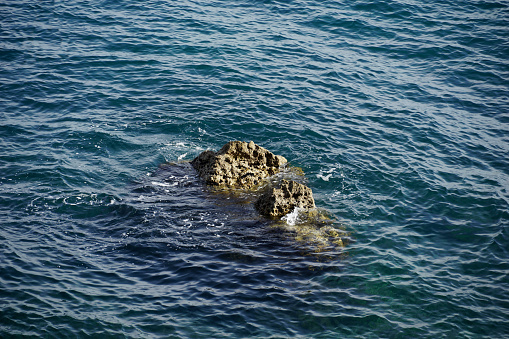 The top of the sea stony rock surrounded by blue wavy sea water