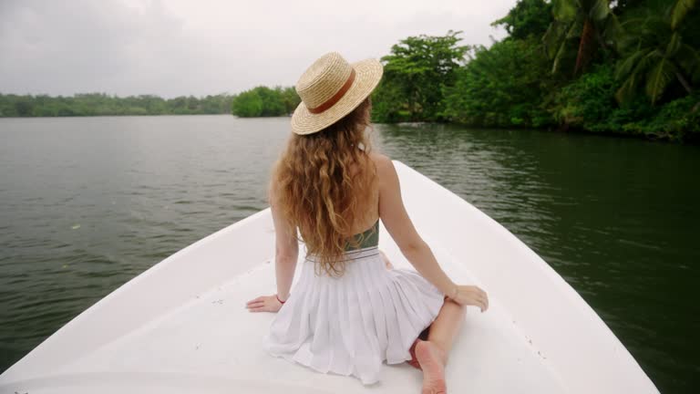 Woman in straw hat enjoys serene river cruise, seated at boat bow, amidst lush tropical greenery. Gentle breeze in hair, tranquil water journey, solo traveler explores natural beauty by river. Slowmo