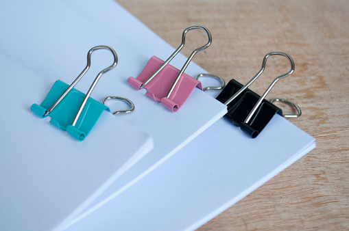 An abundance of silver paper clips on a wooden surface with one colored paperclip in amongst the uniform silver ones.