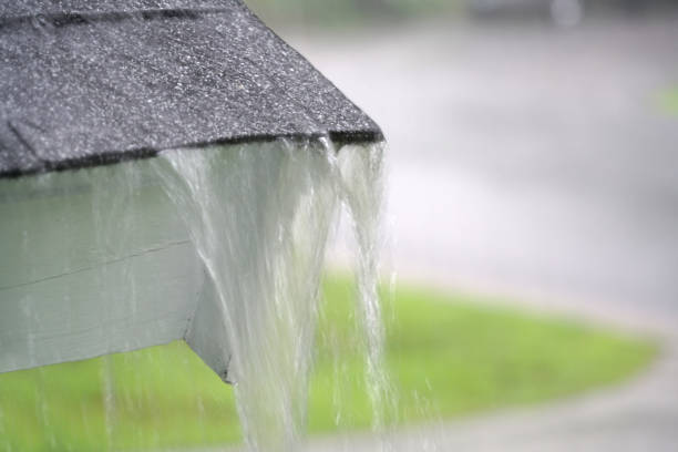 Drenching downpour rain water is splashing over the edge of the tile shingle roof - overflowing out over the overhanging eaves trough aluminum roof gutter system on a suburban residential colonial style house near Rochester, New York State, USA during a torrential mid-summer July storm.