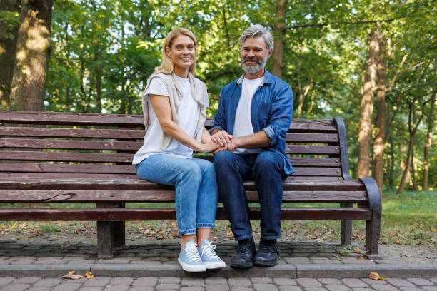 A man and a woman are sitting on a bench in a park. The man is wearing glasses and the woman is wearing a black dress. They are talking to each other