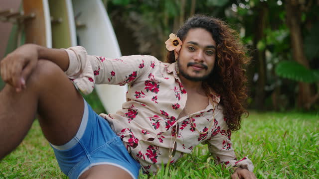 Flamboyant man with eye contacts lounges on grass, posing confidently near surfboards. His style radiates pride, exuding LGBTQ charm and bold fashion choices against a tropical backdrop.