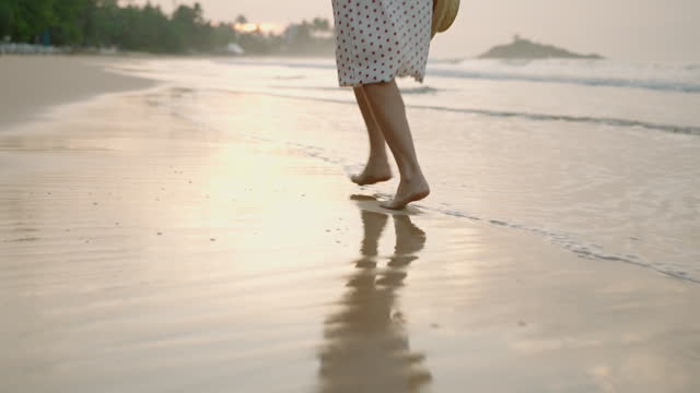 Female feet running barefoot on tropical sandy beach at summer sunset. Slim woman's legs run on sand, leaving footprints. Pretty girl in white dress, carrying straw hat at the seaside surf. Low angle.