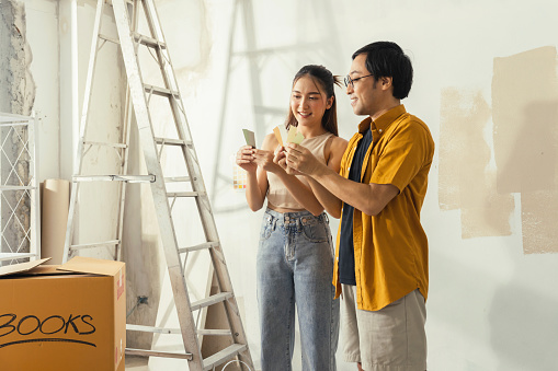 Peaceful Asian Cheerful couple young man and woman smiling sit together on floor painting white wall with roller during renovation house relocation in new apartment Happy couple painting walls