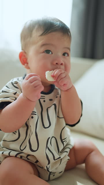 Cute little boy sit sofa in living room and eating rice snack bar.  Asian toddler boy enjoying eating snack at home in living room early in the morning A baby boy eats a rice cracker