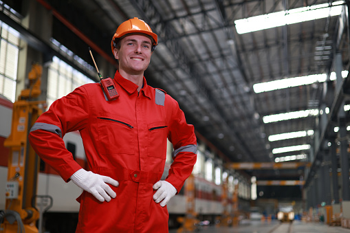 The portrait of a smart railway engineer with a full red safety suit standing on the large maintenance electrical train