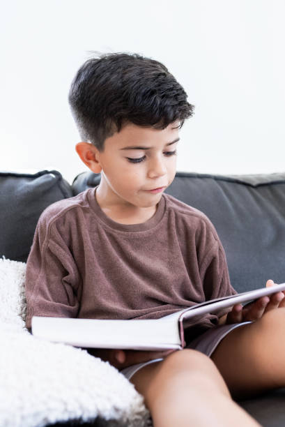 istock Mexican Boy Reading a Book on the Sofa, Learning at Home, Studying. Vertical Photo 2071260500