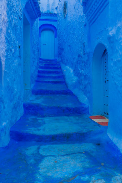 Travel photography from Chefchaouen Morocco, known as the Blue City. The walls, sidwalks, stairs, doors and windows are all painted various shades of vibrant blue hues. The colorful street and alley scenes depict everyday life in a Moroccan tourist town. Handprints and hearts are painted in white on some of the walls and brightly colored robes, curtains and a bakery in the sunlight.