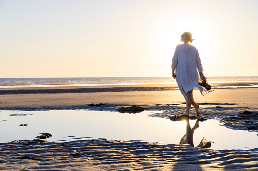 Mature woman walks along empty beach and explores tidal pools at sunset