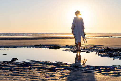 Mature woman walks along empty beach and explores tidal pools at sunset