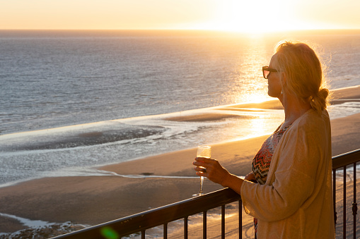 Mature woman holds drink and watches sunset over sea from verandah