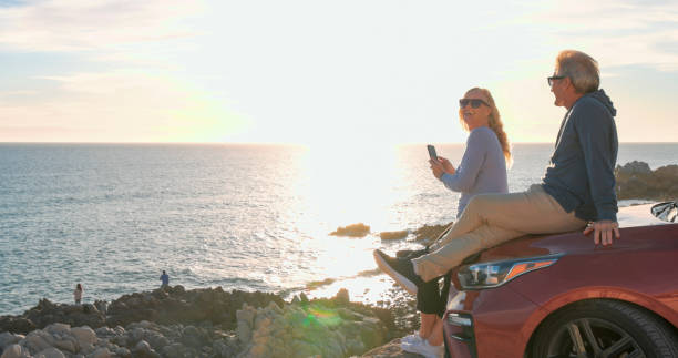 Mature couple pause by sea with vehicle at sunset and woman takes photo