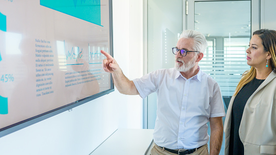 Senior businessman with white hair and beard standing next to an LCD screen with presentation and talking to female colleague in a modern office conference room.