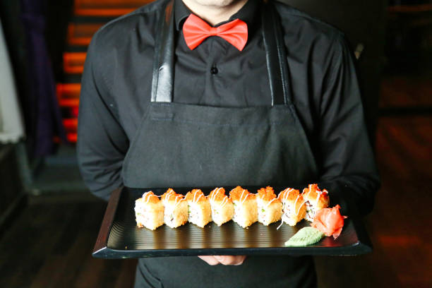 A man wearing an apron holds a tray filled with a variety of sushi.