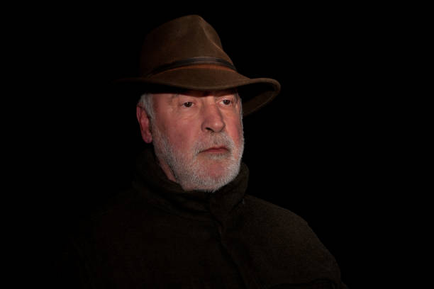 Portrait of a thoughtful looking old hunter with a beard who exudes calm and his face marked by experience, against a black background.