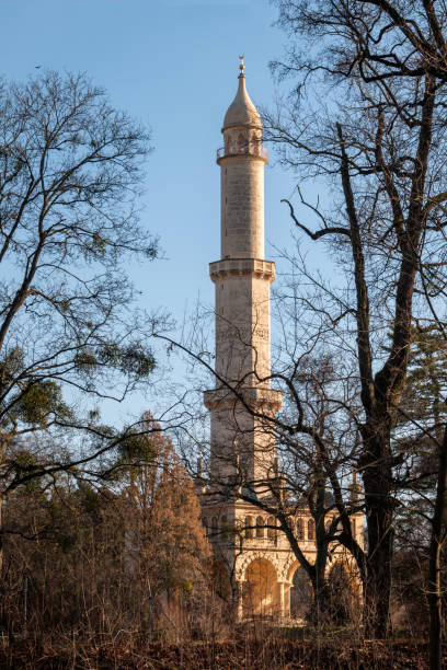 View from the side of river Dyje on a high minaret - viewpoint tower in the park. Surrounded by many leafless trees in the late winter. Bright blue sky. Lednice, Podivin, Morava, Czech republic.