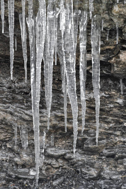 Sharp, crystalline icicles hang precariously from a rugged, icy rock face, covered in a blanket of fresh white snow.