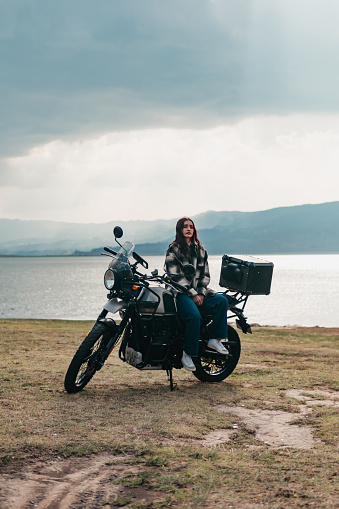 A serene moment captured as a Latina woman sits atop her motorcycle, enjoying the lakeside view with mountains in the distance