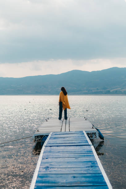 A person stands at the end of a dock, enveloped in tranquility, gazing at the calm waters of a mountain lake