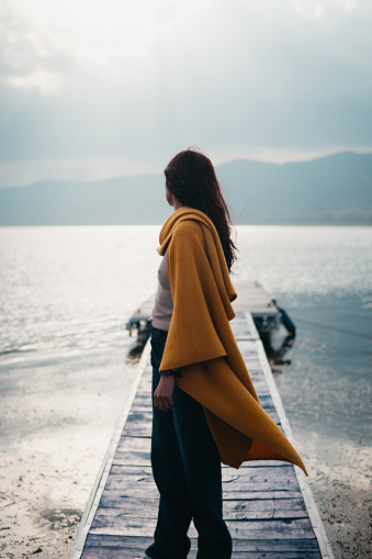 Rear view of a woman wrapped in a golden shawl standing on a dock facing a misty mountain lake