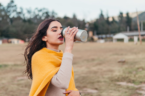 A young Latina woman sipping from a travel mug, wrapped in a cozy yellow sweater, outdoors with nature in the background