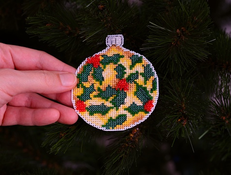 A woman hand holding a handmade cross stitched Christmas ornament against a Christmas tree background. This Christmas ornament made with yellow and green threads, red and green beads by myself.