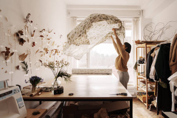 A female tailor holds up a patterned fabric in a well-lit, cozy sewing studio.