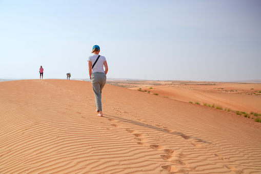 Three senior women walking, one barefoot on Wahiba desert sand in Oman. They are looking to the horizon, only sand, sky and there are some footprints in the sand.