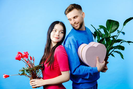 Loving boyfriend holding flowers pot and box and looking to girlfriend with present making romantic surprise.