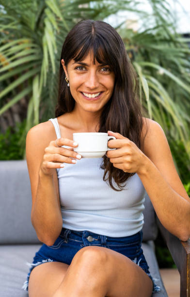 istock A woman is lookintg at camera while sitting on a couch and holding a white cup. She is smiling and she is enjoying her coffee 2071727941