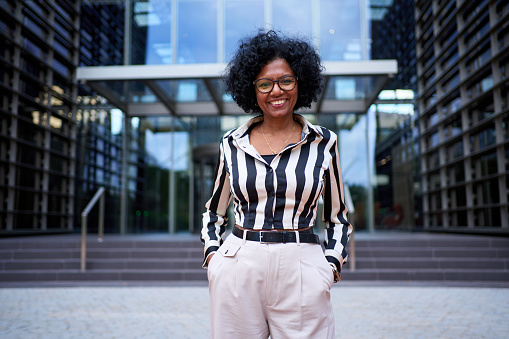 Modern African American business adult woman standing hands in pockets in corporate office area. Strong and empowered black female smiling with curly hair looking joyful at camera outdoor. Copy space