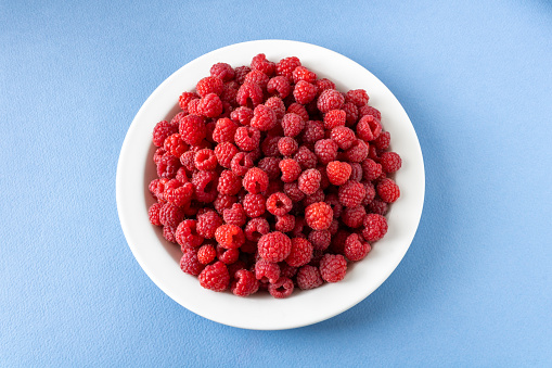 Top down view of a white plate full of raspberries on blue background
