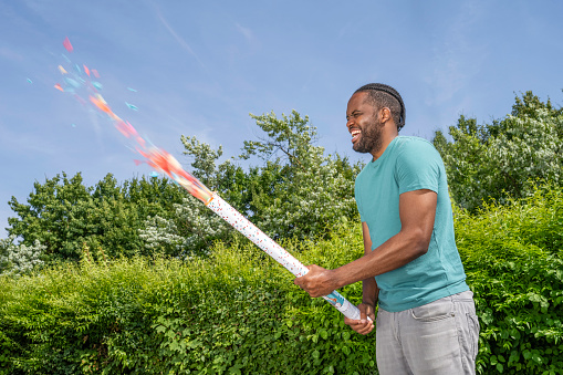 Joyful young african american man shooting party popper with multicolor confetti at festive sunny day, medium shot