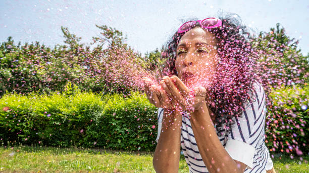 Mid adult hispanic woman blowing pink glitter from palms outdoors at sunny day, medium shot