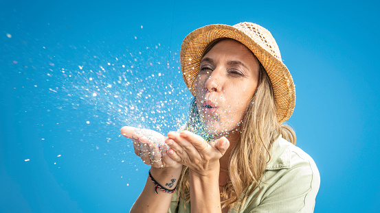 Blonde caucasian woman in sunhat blowing multicolor glitter from palms on blue background, medium shot