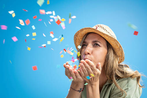 Blonde caucasian woman in sunhat blowing multicolor confetti from palms on blue background, medium shot