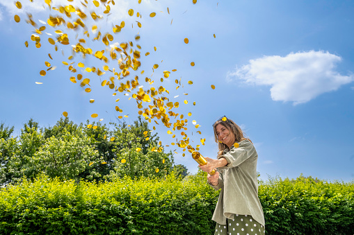 Smiling woman shooting party popper with golden confetti outdoors at sunny day, medium shot