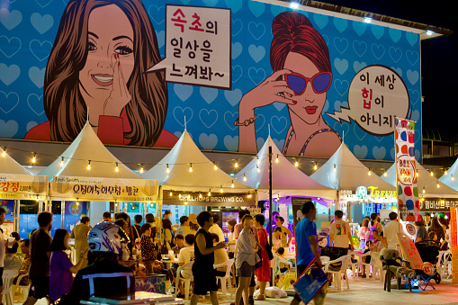 Sokcho City, South Korea - July 30, 2019: An active night market on Sokcho Beach, under a large building with murals and white popup tents illuminated by overhead lights, as people bustle around the market.