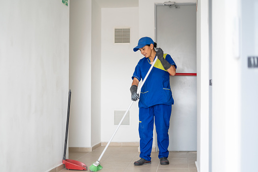 The woman from the cleaning and general maintenance services uses a broom and dustpan to clean and collect the garbage that is in the hallways and corridors of the apartment area
