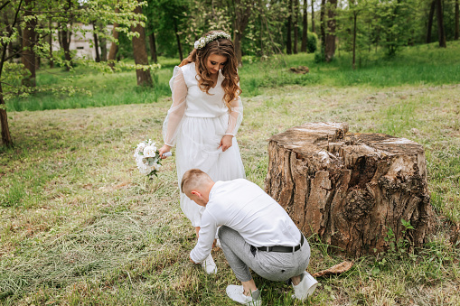 bridal walk in the forest. The groom is dressed in a white shirt and gray pants, helping the bride to tie her shoelaces