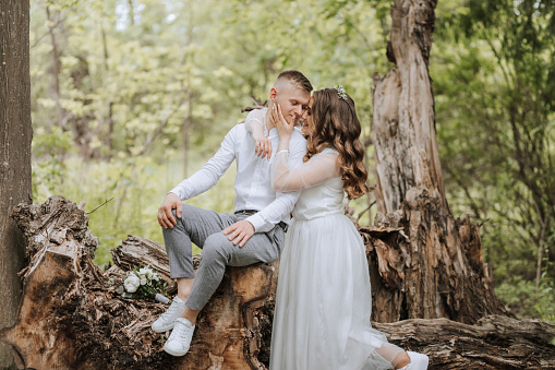 Gentle embrace of the bride and groom in the forest. The groom is dressed in a white shirt and gray pants, the bride is in a light white dress