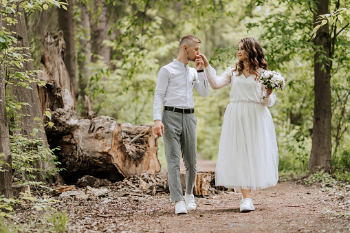 Wedding walk in the forest. The groom kisses the bride's hand and they go in front of the big trees looking at each other