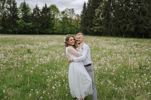 the bride and groom walk in a field with dandelions on their wedding day. The groom hugs the bride, smiles and kisses