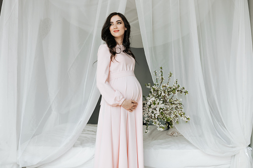 A beautiful pregnant woman in a pink dress holds her hands on her stomach. A loving mother is waiting for the birth of a child. Concept of pregnancy, motherhood, preparation and waiting.